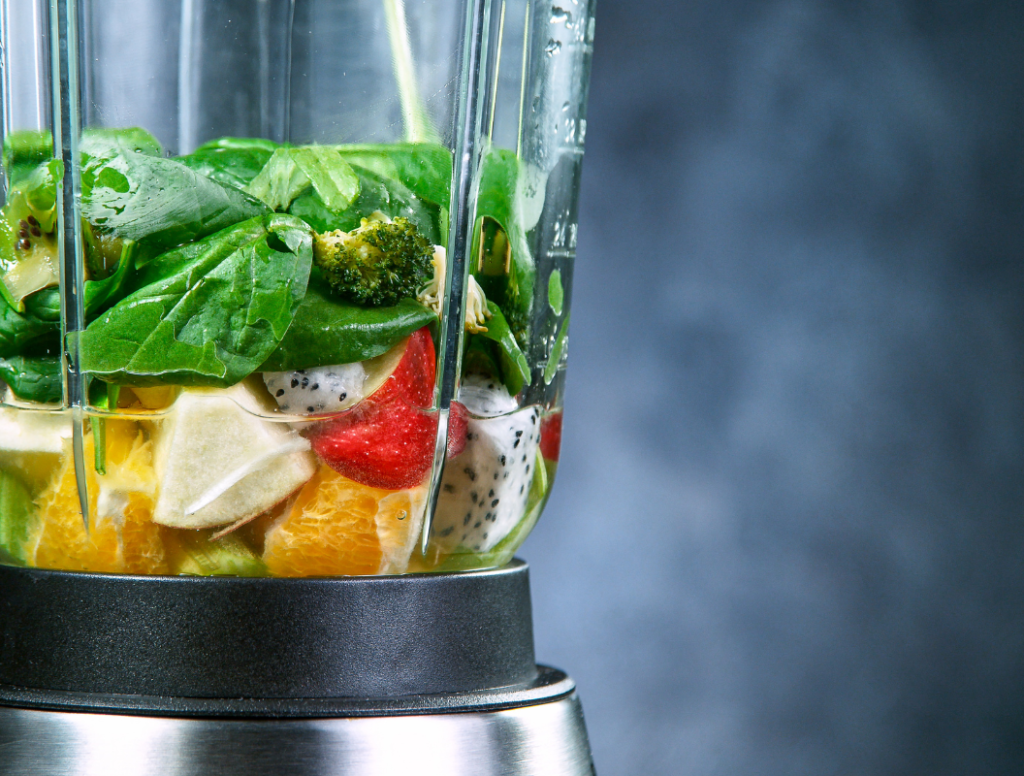 Ingredients for a green smoothie recipes in a blender, including spinach, broccoli, strawberries, orange slices